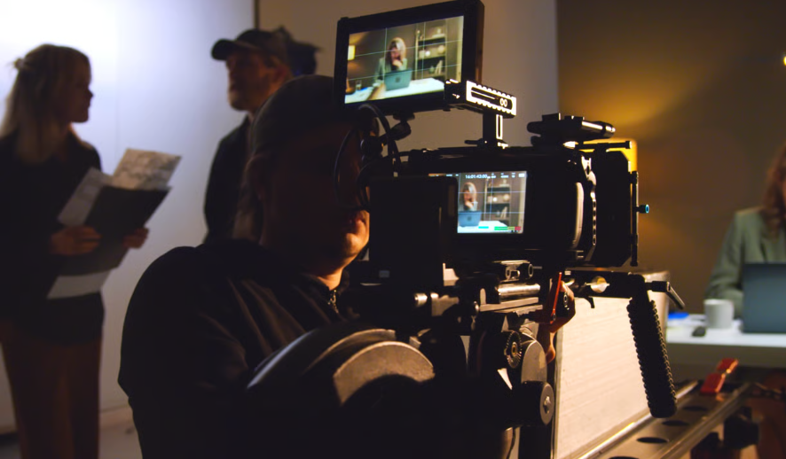 Camera operator films a woman on a monitor during a dimly lit indoor production.