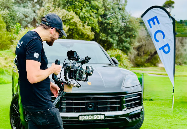 Man filming a dark SUV with a professional camera beside an “ECP” flag outdoors.