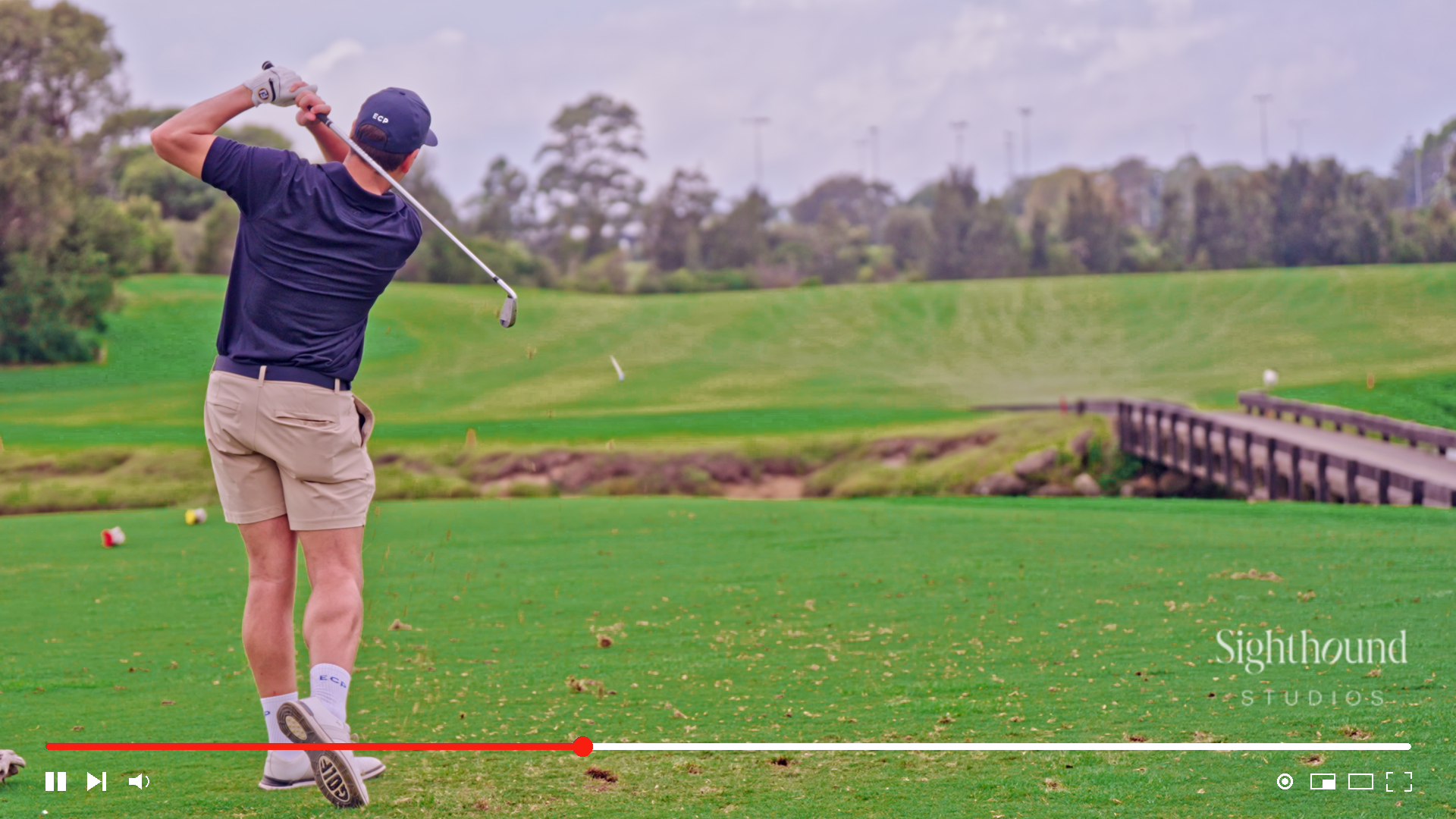 Golfer in mid-swing on a green course with a bridge, trees, and video controls visible.
